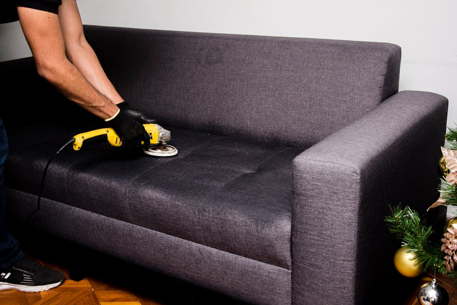 A person wearing black gloves is using a small handheld electric polisher to deep clean the dark grey fabric upholstery of a sofa in a living room. The sofa has a modern design with a high backrest and wide armrests, positioned on a wooden parquet floor. The room is well-lit with natural light and features a white wall in the background, along with a decorated Christmas tree partially visible on the right side. This scene exemplifies surface cleaning and maintenance for domestic furniture, with the focus on achieving a hygienic and refreshed appearance. Sutton Cleaners provides professional upholstery cleaning services to ensure furniture is thoroughly sanitised and looks its best.
