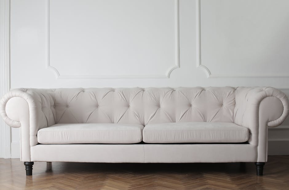 A clean and elegant beige upholstered sofa with a tufted design positioned against a white paneled wall in a well-lit living room. The sofa features rolled arms and dark wooden legs, and the surface appears spotless and freshly maintained, reflecting thorough surface cleaning. The hardwood floor beneath is smooth and polished, adding to the room's tidy appearance. This setting exemplifies domestic cleaning and surface sanitisation performed by Sutton Cleaners, highlighting their expertise in deep cleaning for upholstery and maintaining hygienic living spaces.
