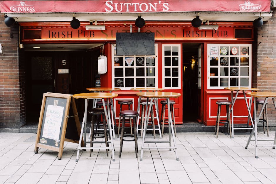 The exterior of Sutton's Irish Pub features a red facade with white window frames and door, located on a paved sidewalk. There are three high wooden tables with matching stools positioned in front of 