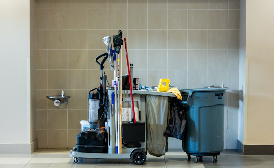 An industrial cleaning trolley positioned against a beige tiled wall in a commercial environment, containing various cleaning tools including mops, cloths, spray bottles, and a vacuum cleaner. To the right, there is a large blue waste bin with a lid, and a small stainless steel sink mounted on the wall to the left. The surface areas appear clean and organized, with the trolley holding cleaning equipment ready for use. The lighting is bright and neutral, emphasizing the cleanliness of the space, which is likely part of an office or commercial facility. The scene reflects professional cleaning service standards provided by Sutton Cleaners, suitable for maintaining hygiene and sanitation in business environments.